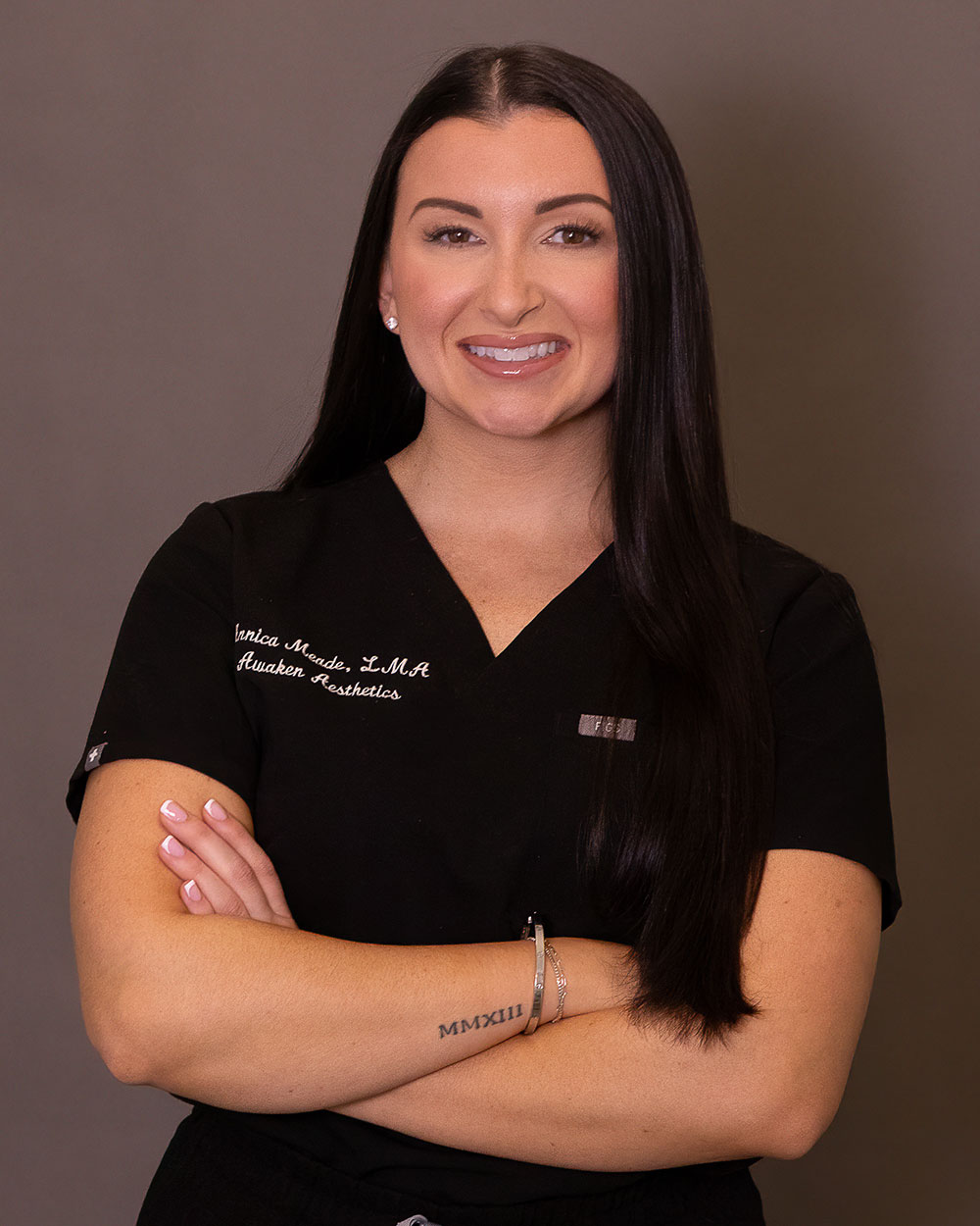 Smiling woman in black medical scrubs with long dark hair, arms crossed, neutral background