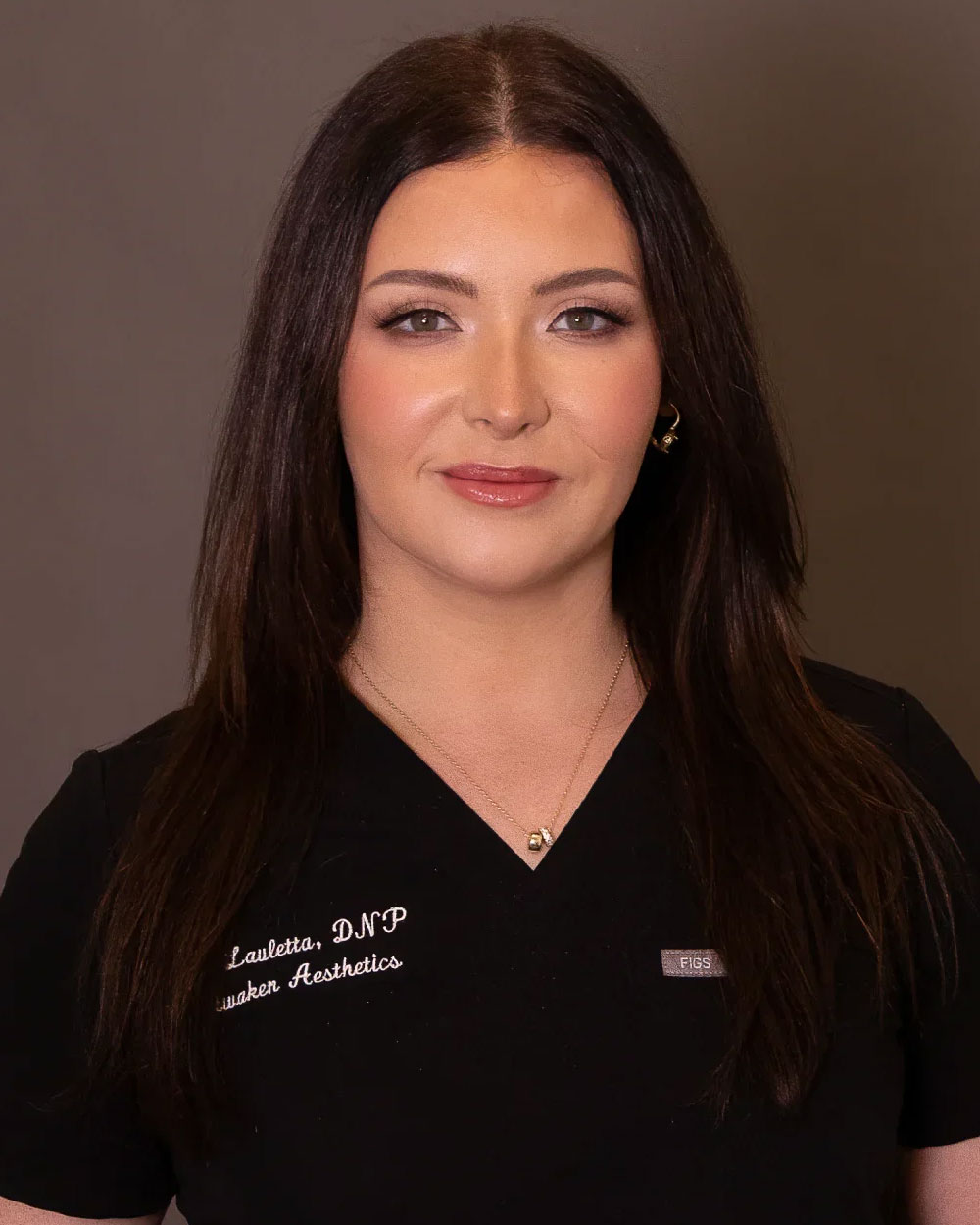 Headshot of a woman in black medical scrubs with long dark hair and light makeup against neutral background