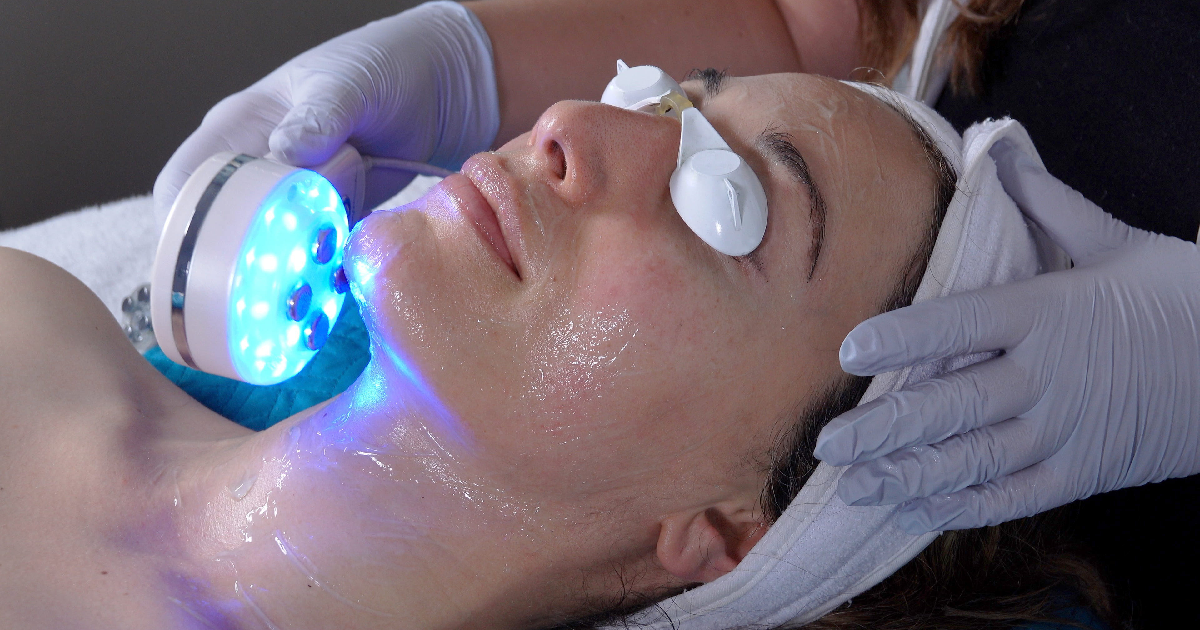 A woman receiving blue LED light facial treatment on chin and neck, esthetician wearing gloves and using handheld device.