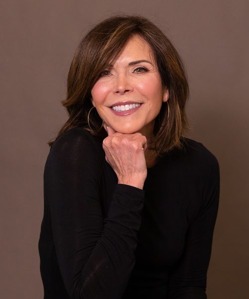 Smiling woman with brown hair and hoop earrings in a black top, resting chin on hand against a neutral backdrop.