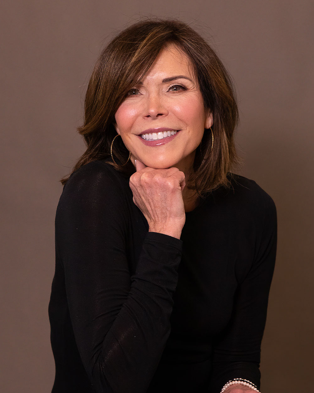 Smiling woman with brown hair and hoop earrings in a black top, resting chin on hand against a neutral backdrop.