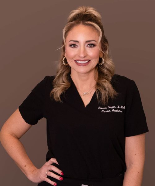 Smiling woman in black medical scrubs with gold hoop earrings, hand on hip, against a brown studio background.