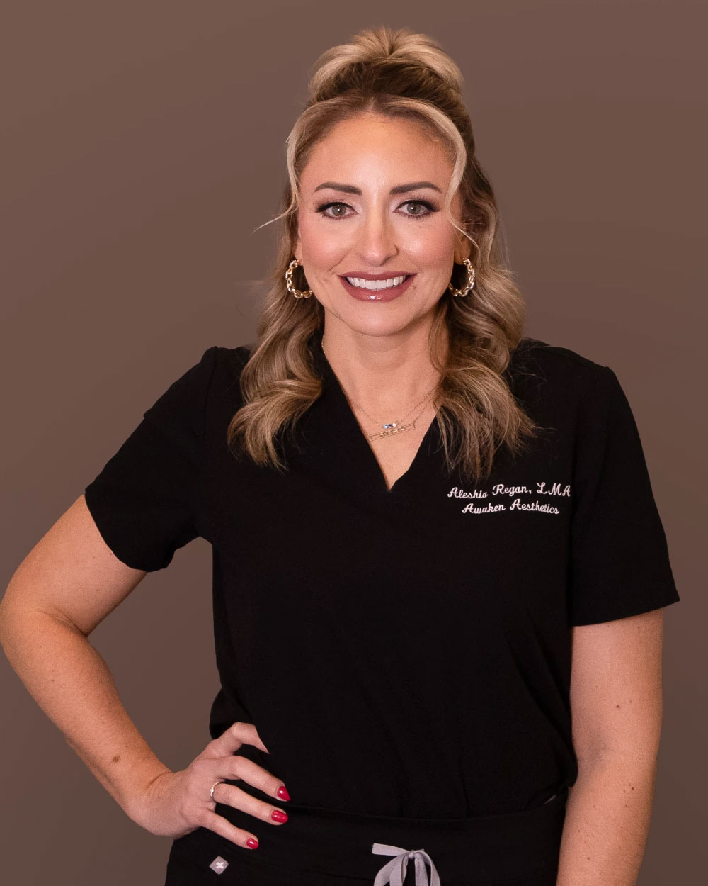 Smiling woman in black medical scrubs with gold hoop earrings, hand on hip, against a brown studio background.