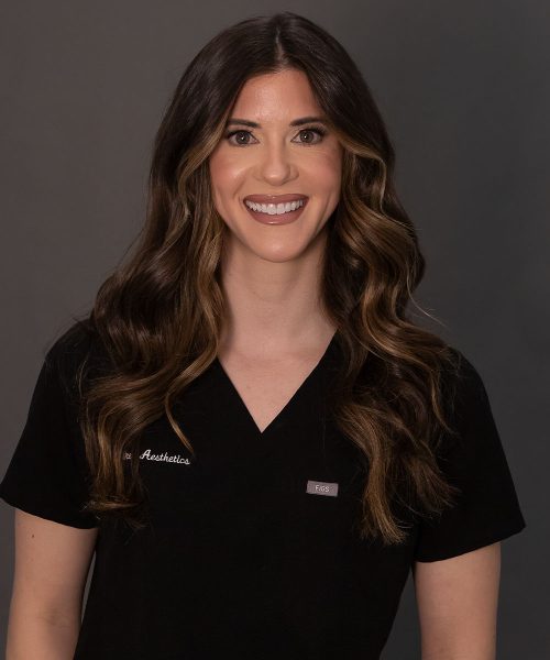 Smiling woman in black medical scrubs with long wavy brown hair against a neutral gray background