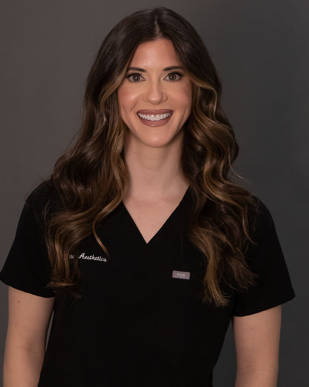 Smiling woman in black medical scrubs with long wavy brown hair against a neutral gray background