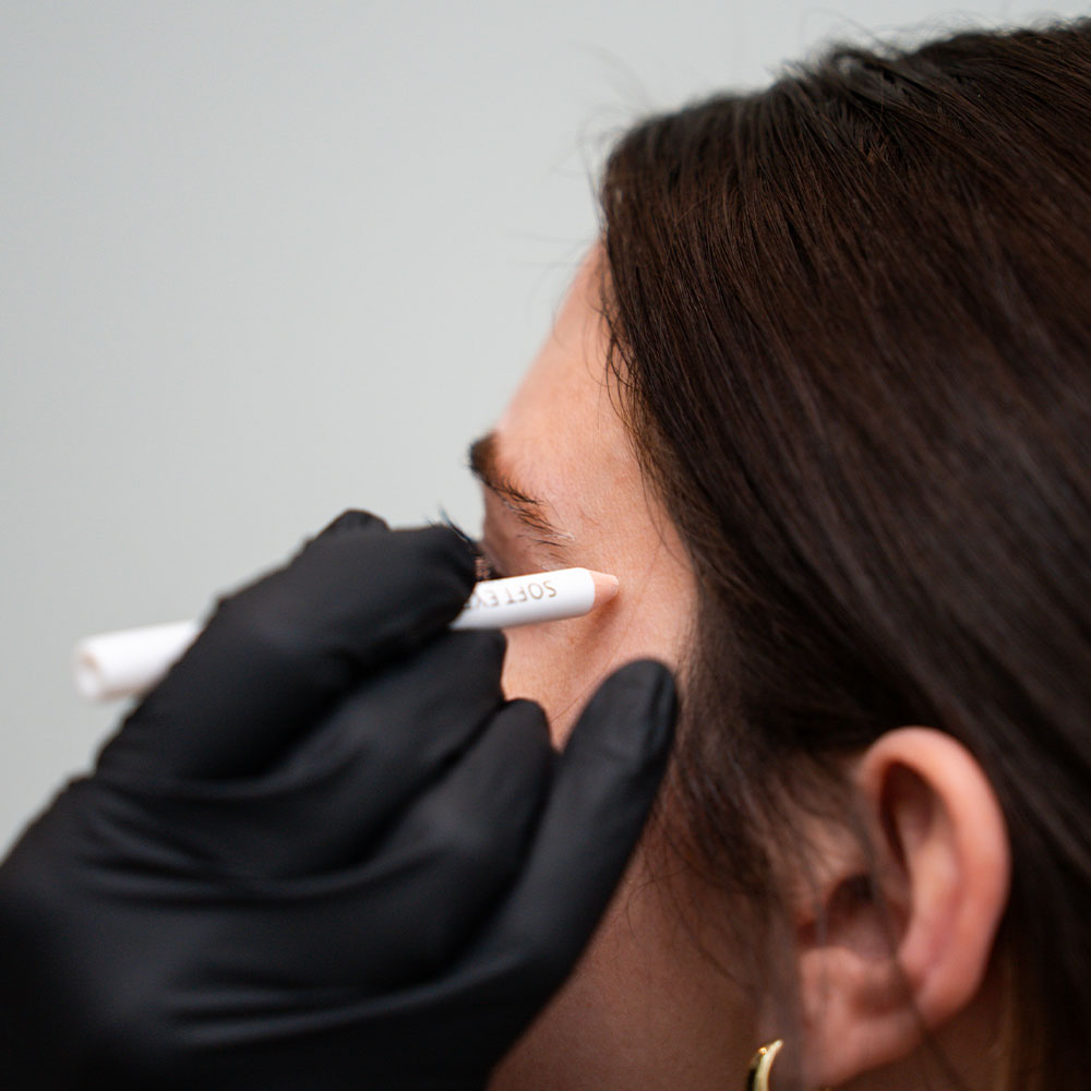 Esthetician marking a woman's eyebrow with a white pencil while wearing black gloves for cosmetic treatment