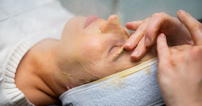 A woman receiving a yellow facial mask at a spa as an esthetician gently applies it near her eye.