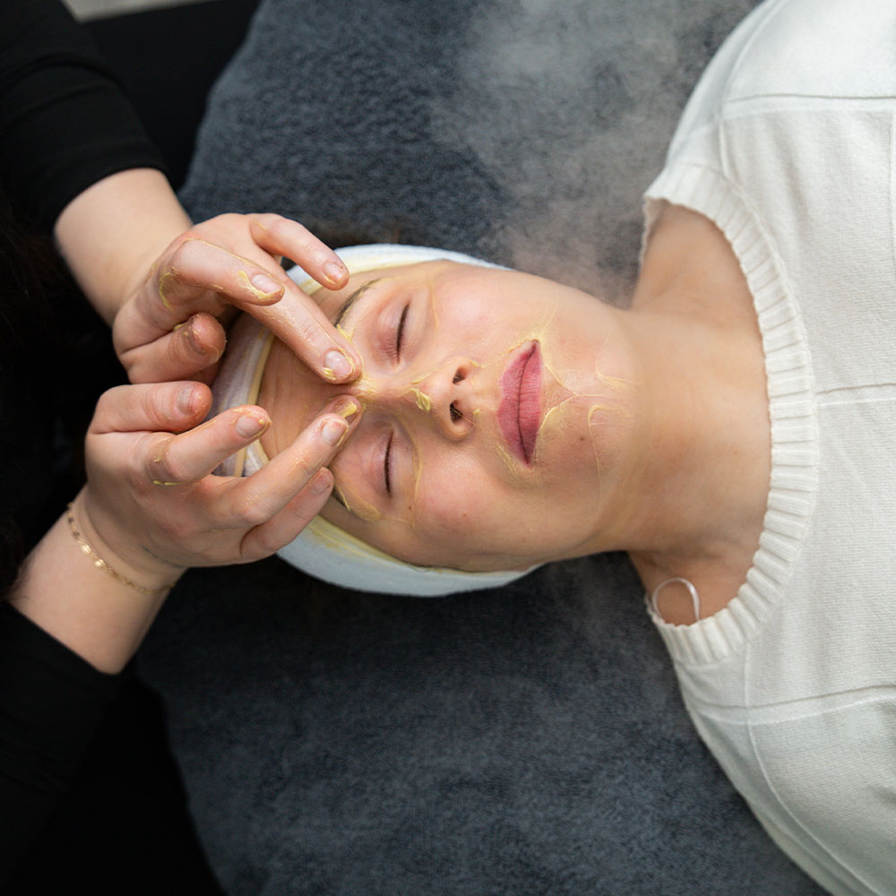 A woman receiving a yellow facial mask and gentle eyebrow massage while lying back at a spa.