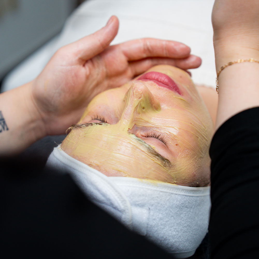 A person receiving a yellow facial mask while an esthetician's hands gently touch their face.