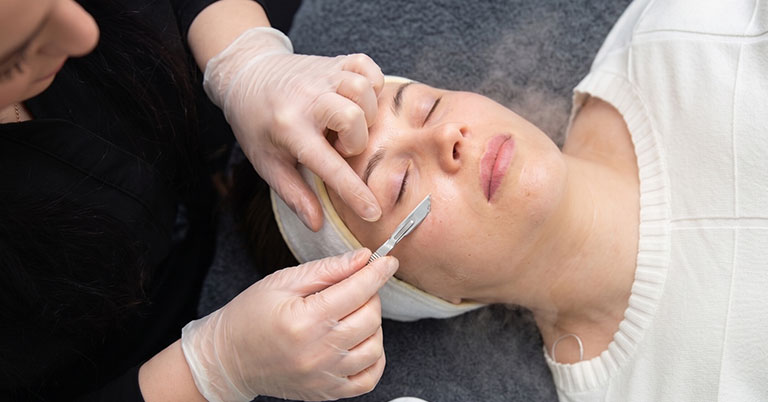 Esthetician performing dermaplaning on a woman's face with a straight razor during a facial treatment