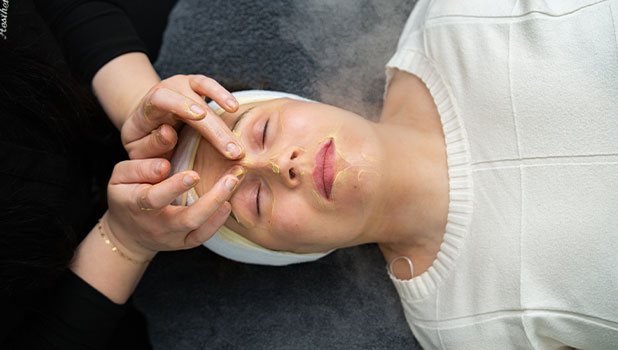 A woman receiving a facial treatment as an esthetician applies a nourishing mask around her eyes and nose.