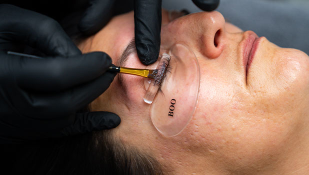 Close-up of a woman getting eyelash extensions as a technician applies adhesive with a brush and under-eye gel pad.