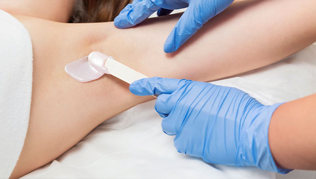 A woman receiving underarm waxing hair removal while technician in blue gloves applies wax