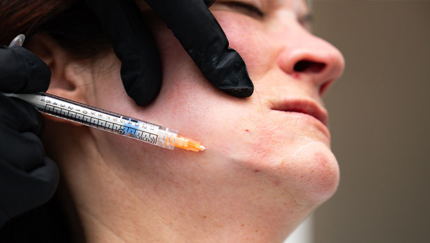 Woman receiving cosmetic cheek injection with syringe from clinician wearing black gloves