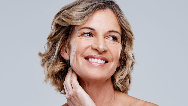 Smiling mature woman with short wavy blonde hair touching neck in skincare beauty portrait on light gray background.
