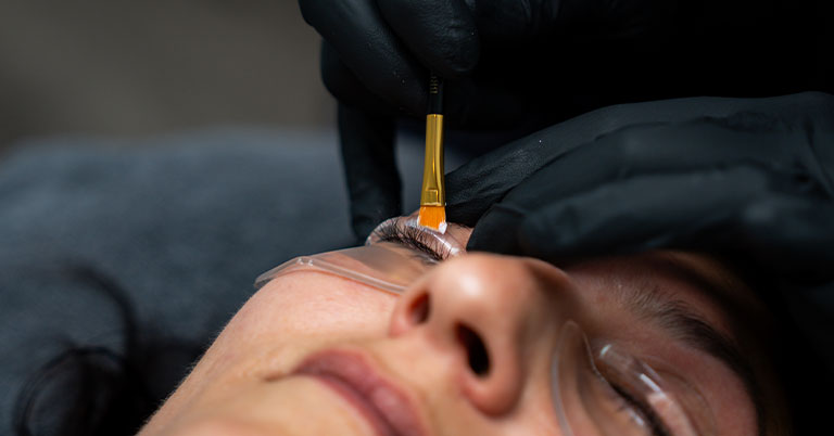 Close-up of a woman receiving eyelash extension application with brush and under-eye gel pads.