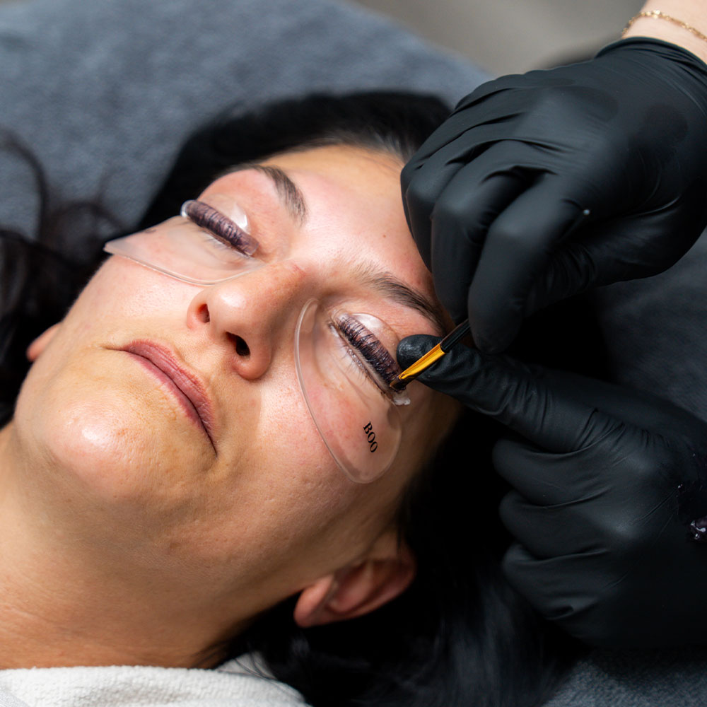 A woman reclines with under-eye gel pads while a technician in black gloves applies eyelash extensions.