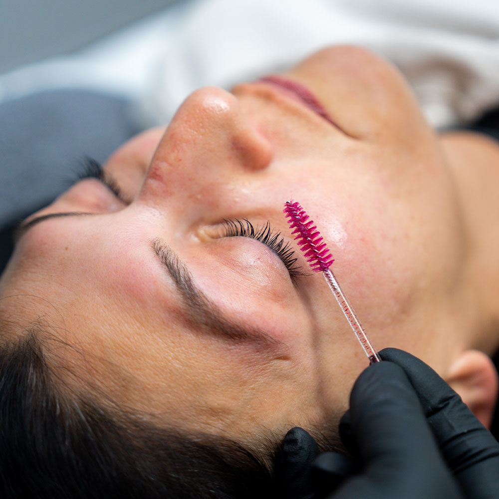 A woman with eyes closed receiving eyelash brushing with a pink spoolie from a gloved technician.
