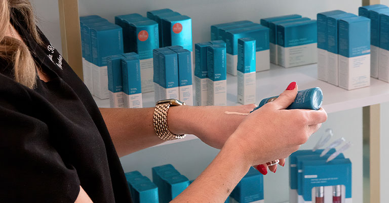A woman testing a blue skincare lotion on her hand in front of shelves of matching blue product boxes.