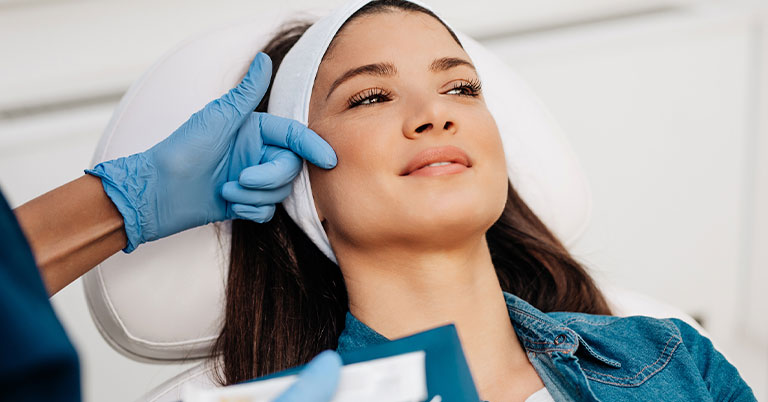 A woman reclining in a skincare clinic while an esthetician in blue gloves examines her face.