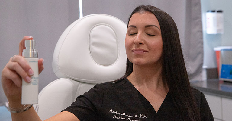 A woman in a spa uniform sprays facial mist with eyes closed in a skincare clinic treatment room