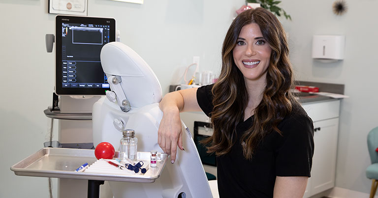 Smiling woman in dental clinic by exam chair with tray of instruments and monitor.