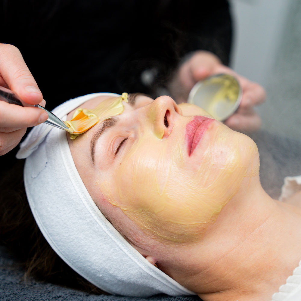 A woman receiving a yellow facial mask at a spa, eyes closed as an esthetician applies it with a spatula