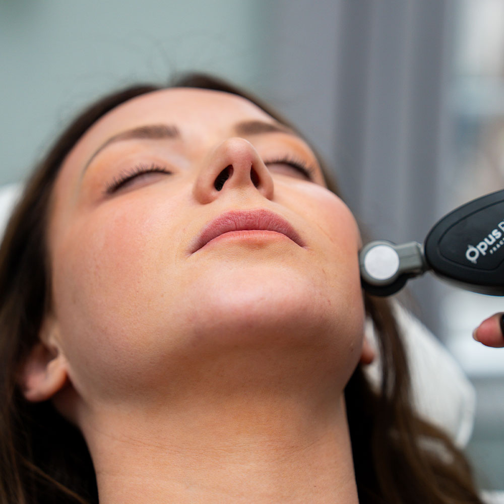 A woman reclined with eyes closed receiving a handheld cosmetic skin treatment along her jawline
