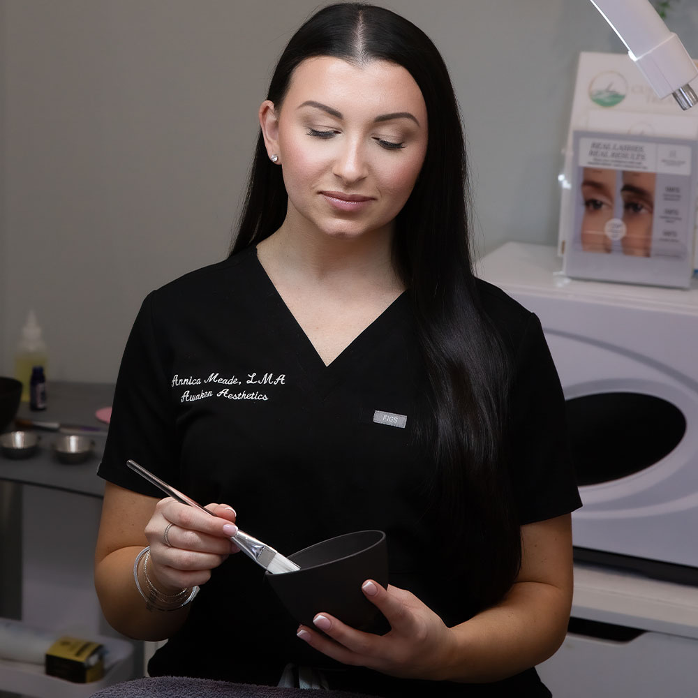 A woman esthetician in black scrubs mixing a facial mask in a bowl with a brush in a skincare treatment room.