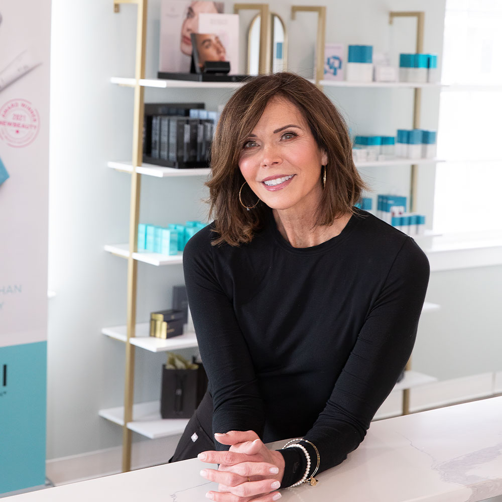Smiling woman leaning on a clinic counter with skincare product shelves in the background.