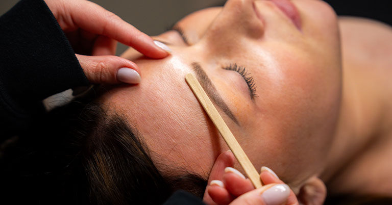A woman receiving eyebrow waxing and brow shaping with a wooden spatula by an esthetician.