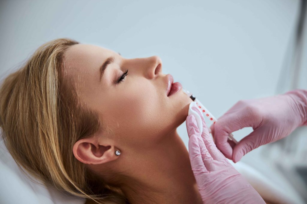 Woman receiving dermal filler chin injection from clinician in pink gloves