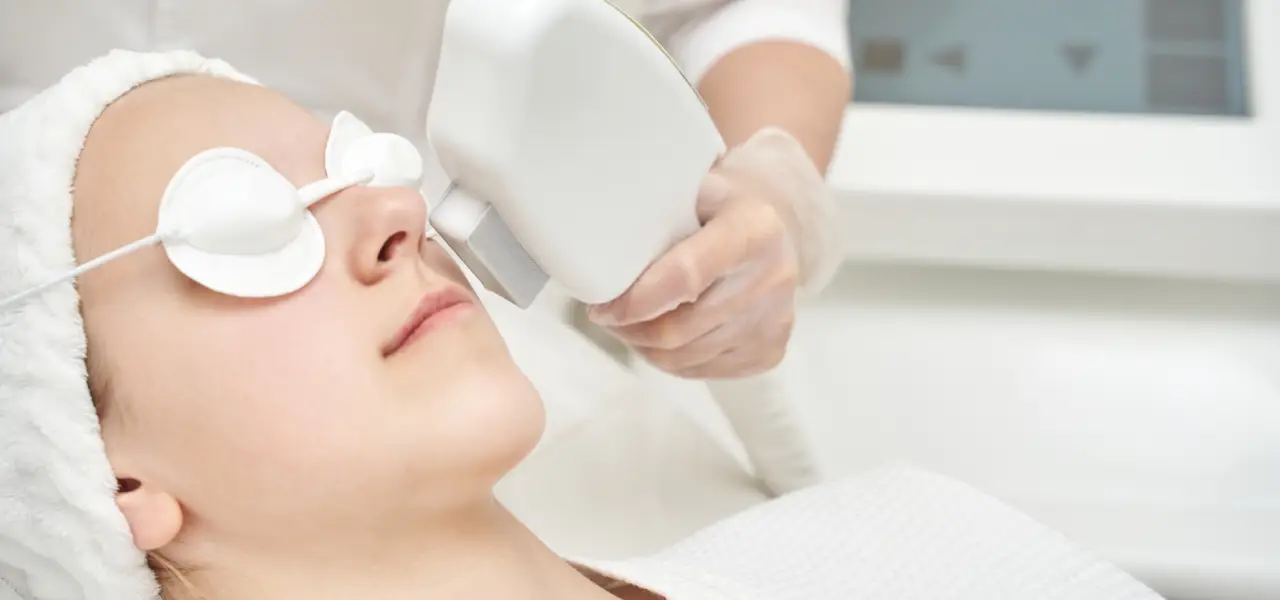 Woman receiving facial laser/IPL treatment with protective eye pads from technician in clinic
