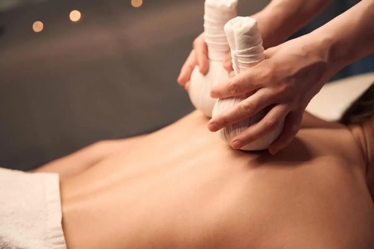 A person receiving a hot herbal compress massage on their bare back in a relaxing spa setting