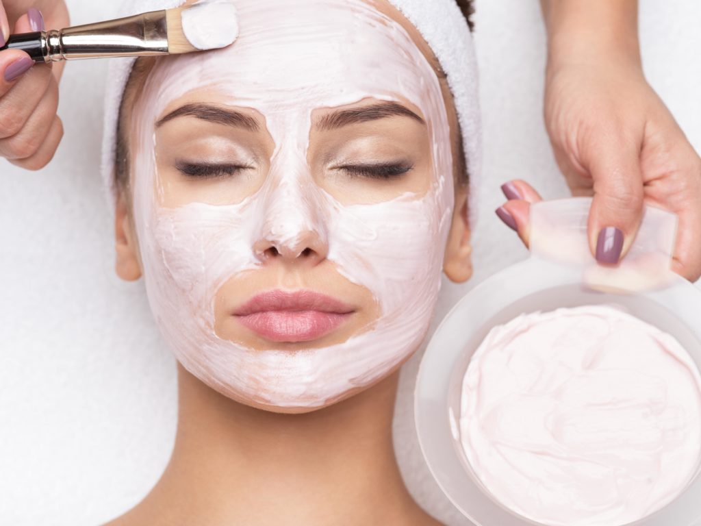 Woman with eyes closed getting a pink facial mask applied with a brush, bowl of cream held nearby.