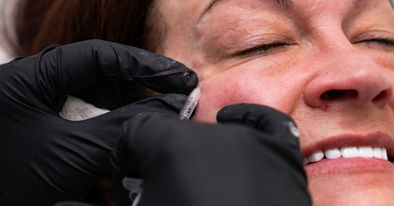 Close-up of a woman receiving a cosmetic facial injection to the cheek from a clinician wearing black gloves