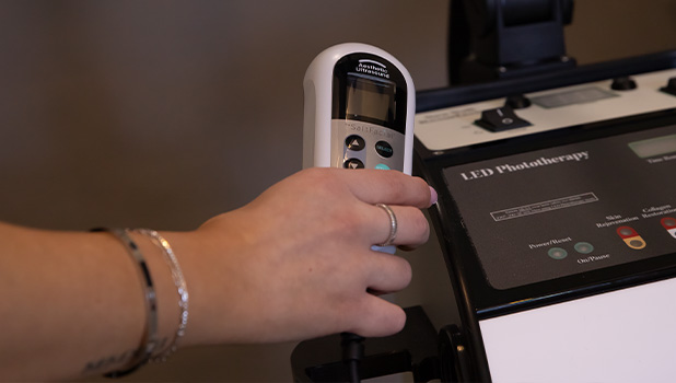 A person's hand holding a handheld controller for an LED phototherapy machine, wearing bracelets and rings.