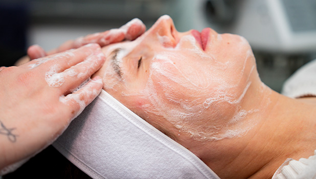 A woman receiving a foamy facial cleansing treatment while wearing a white headband.