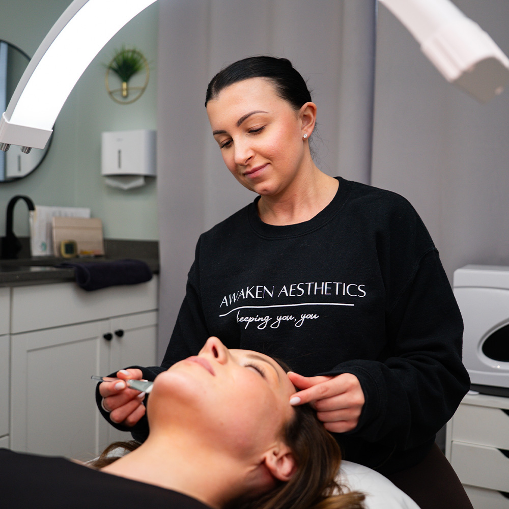 Woman esthetician in Awaken Aesthetics sweatshirt performing facial treatment on a client in a treatment room