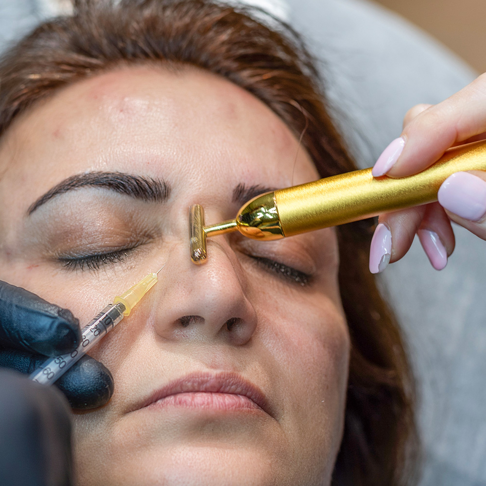 Close up of a woman receiving a cosmetic injection near the nose while a gold T-bar facial massager rests on her forehead.
