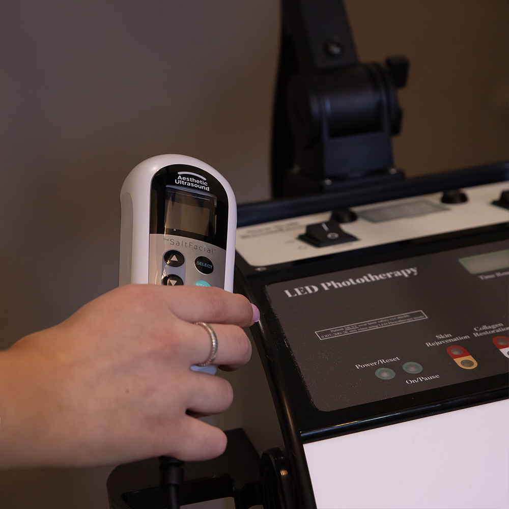Hand holding an aesthetic ultrasound device next to an LED phototherapy machine for skin treatment in a clinic.