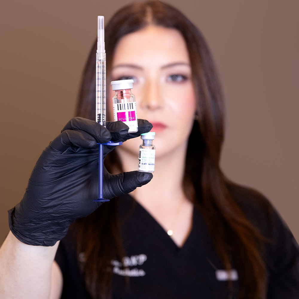 Gloved hand holding syringe and two cosmetic vials in focus, a woman aesthetician blurred in background